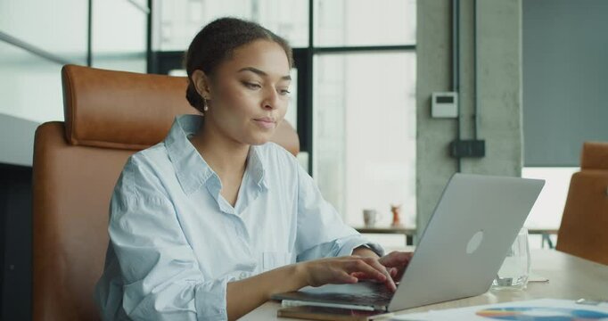 A focused young woman working on a laptop in a modern office environment, interacting with a smartphone and analyzing documents on a desk with bright natural lighting