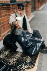 Full length vertical portrait of older homeless man sitting on urban street with belongings. Sad aged tramp seeking assistance, holding disposable cup and begging for alms. Concept of poor retirement.