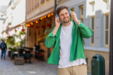 Fototapeta premium Handsome young man tourist in wireless headphones listening favorite energetic disco music outdoors. Happy Caucasian male traveler walking passes by urban sunny city street. Town lifestyles