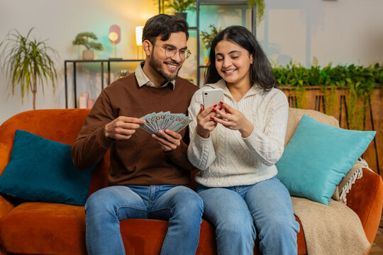 Happy Indian couple holding money cash and smartphone planning discussing investment together sitting on sofa in living room at home. Successful diverse family on couch in apartment.