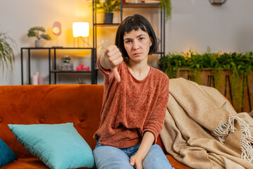 Obraz premium Portrait of upset Caucasian woman showing double thumbs down sign gesture, expressing discontent, disapproval dissatisfied bad work dislike sitting in living room at home. Adult girl sitting on couch