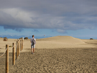 A man tourist walking along a sand desert dune at the background of cloudy sky in Maspalomas, Gran Canaria