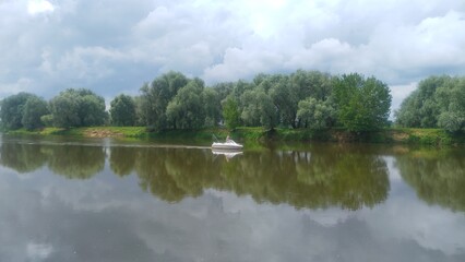 A motorized plastic boat floats on the river, and fishermen catch predatory fish by trolling. Branches of willows growing along the banks lean over the water. The summer sun breaks through the clouds