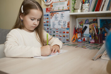 A young girl sits at a desk in a colorful and organized learning space, writing attentively. The setting reflects creativity with artwork and school supplies surrounding her.