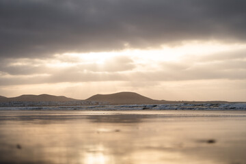 Low angle of sunset over calm ocean reflecting distant hills and cloudy sky