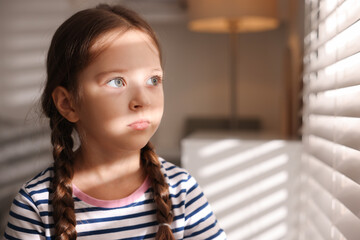 Orphanage concept. Sad girl and looking through window blinds indoors