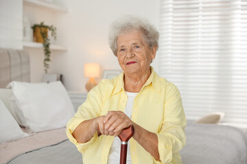 Senior woman with walking cane sitting on bed at home