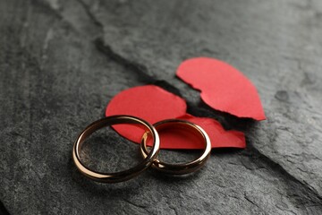 Halves of torn red paper heart and wedding rings on dark grey table. Broken heart