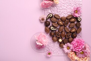 Heart made with delicious chocolate candies and beautiful flowers on pink background, flat lay. Space for text