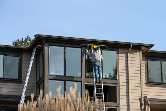 Workman high up on an aluminum extension ladder against a large two-story house, window replacement construction project, outside on a sunny winter day
