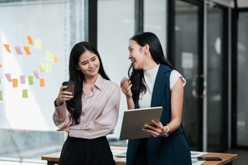 Two businesswomen engaged in a lively discussion in a contemporary office environment, holding a tablet and coffee cup.