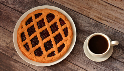 A whole cherry pie on a large ceramic plate and a cup of hot black coffee on a wooden table. Top view, flat lay.