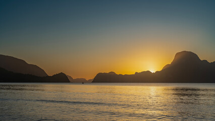 Beautiful tropical sunset. The blue sky is tinged with orange near the horizon. The setting sun is shining from behind the mountains. Reflection on the calm ocean surface. Long exposure. Philippines. 