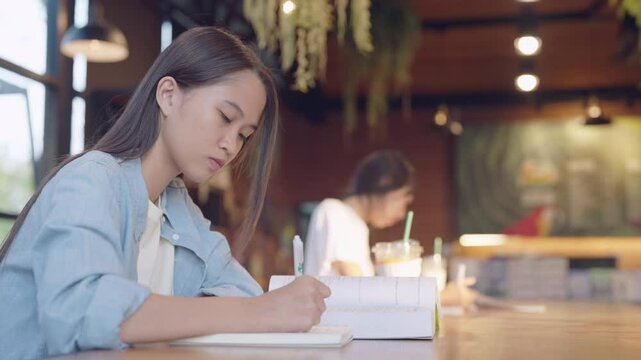 A woman is sitting at a table with a notebook and pen. She is writing something down. The scene is set in a cafe with other people around