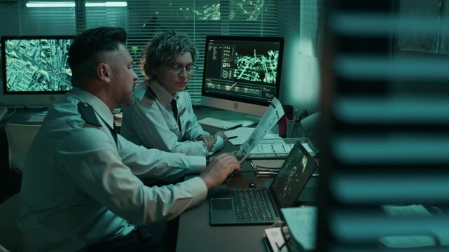 Medium shot of two mature Caucasian male and female military officers in uniform shirts with shoulder patches sitting in command center, reading dossier, discussing secret intelligence information