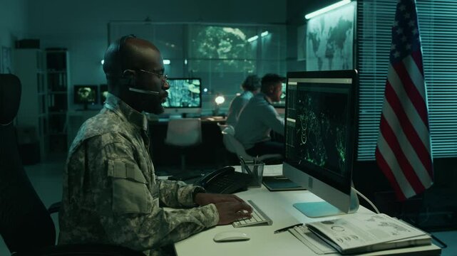 Medium side shot of African American male military officer in camouflage uniform operating surveillance satellite on computer, while working in operations center, USA flag in background