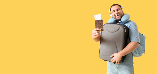 Young happy man with suitcase, headphones, passport and travel pillow on yellow background
