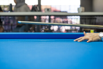 A hand strikes a billiard ball with precision using a cue stick during an intense pool game on a blue table, highlighting focus, skill, and competitive gameplay in an indoor setting.