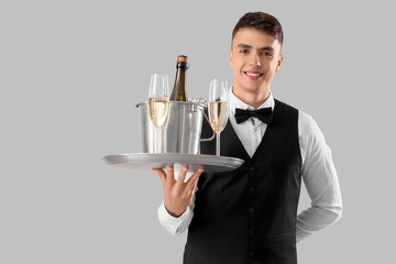 Young male waiter with champagne and ice bucket on tray against grey background
