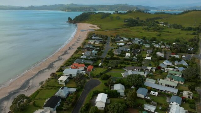 Coastal town nestled beside a sandy beach. Houses line the shore, showcasing diverse architecture. Aerial view. SIMPSON BEACH, WHITIANGA, COROMANDEL PENINSULA, NEW ZEALAND