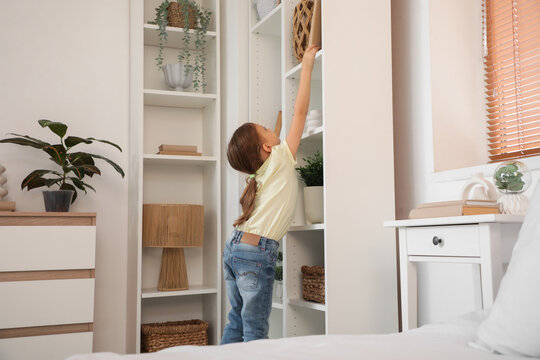 Cute little girl taking book from high shelf at home. Child in danger