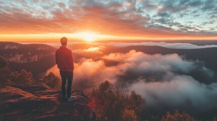 Obraz premium A person stands on a cliff, gazing at a vibrant sunset over a misty landscape.