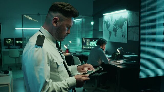 Medium side shot of focused Caucasian army lieutenant in uniform shirt with shoulder patches standing in operations center, using tablet computer with digital satellite maps to plan troop movements