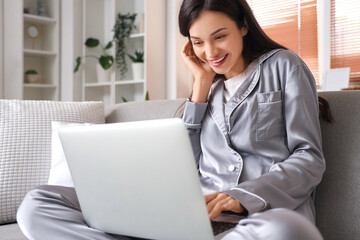 Young woman in grey pajamas working with laptop on sofa at home