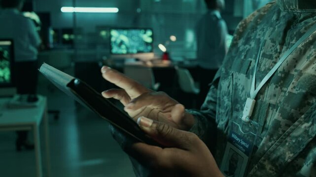 Tilting closeup of hands and face of young African American army field commander in camouflage uniform standing in operations center, using tablet computer while transmitting enemy coordinates