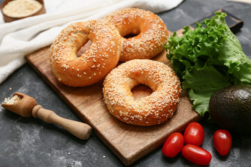 Wooden board of tasty bagels with sesame seeds and fresh vegetables on black background