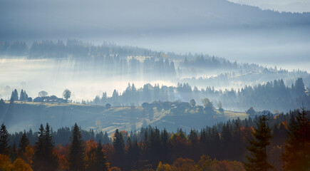 Naklejka premium Morning sunlight streams through mist, illuminating serene mountain village nestled among forested hills. Light creates ethereal rays that gently touch autumn foliage and houses scattered on slopes.