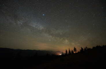 Breathtaking night sky displays dense field of stars and Milky Way over tranquil mountain landscape. Silhouetted trees and distant mountain peaks faintly illuminated by glow from below horizon.