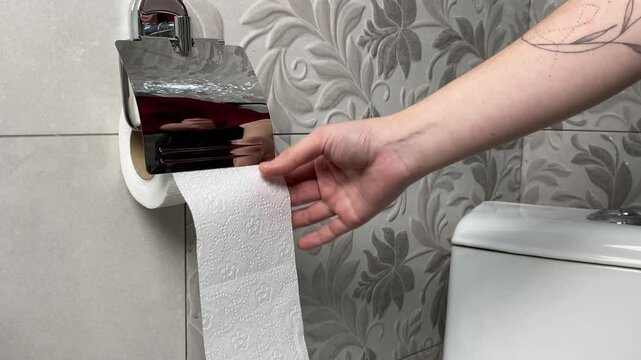 A woman's hand unrolls a roll of toilet paper in a bathroom.