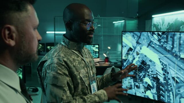 Medium shot of African American male army officer and Caucasian operation center strategist talking while analyzing surveillance footage of enemy industrial objects, choosing bomb targets