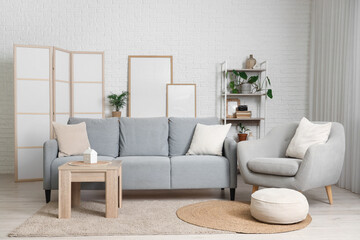 Cozy sofa, armchair, coffee table and blank posters near white brick wall in living room