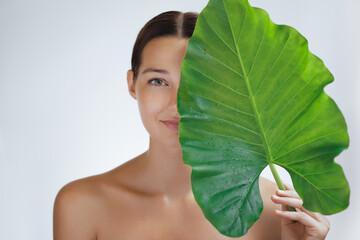 A beautiful woman with glowing skin holds a large green leaf, partially covering her face. The image represents natural beauty, eco-friendly skincare, and wellness, perfect for organic beauty