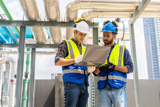 Collaboration, Two young construction workers discuss plans while reviewing blueprints on a rooftop. They wear safety gear and oversee intricate piping systems amidst a bustling urban backdrop,