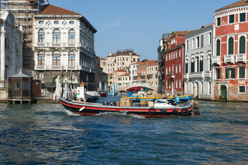 A boat carrying packages navigates a Venetian canal, surrounded by historic buildings and a bridge,...