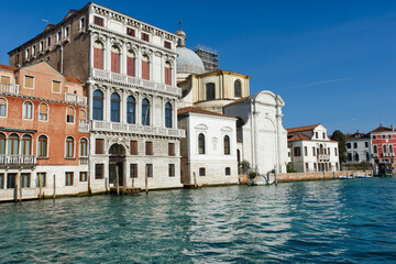 Obraz premium A charming Venetian scene with vibrant buildings and a domed church reflecting in the serene canal under a clear blue sky, capturing Venice's historic and architectural beauty.