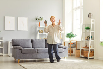 Happy elderly woman dancing joyfully to music and holding phone at home living room, celebrating...