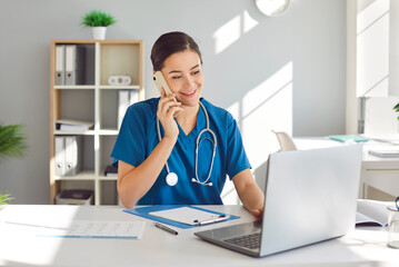 Happy friendly young woman doctor, nurse, medical worker, reception specialist in blue uniform with stethoscope sitting at desk, talking to patient on phone and checking schedule information on laptop
