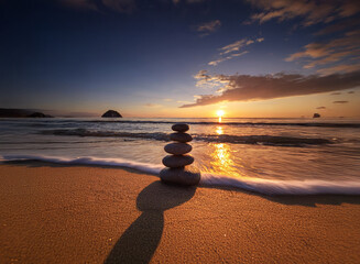 Serene sunrise over a tranquil beach.  A carefully balanced stack of stones stands on the golden sand, casting a long shadow as gentle waves lap the shore.  The vibrant colors of dawn paint the sky.