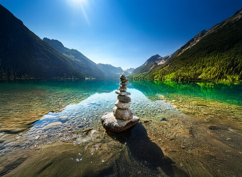Serene mountain lake scene with a carefully balanced rock cairn at the water's edge.  Clear, turquoise water reflects the sunlit peaks. Peaceful, idyllic landscape. - Powered by Adobe
