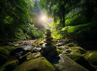 Sunbeams pierce a lush, green rainforest, illuminating a carefully balanced stone cairn beside a moss-covered stream. Serene nature scene.