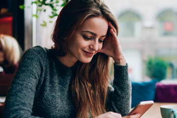 Smiling successful female student watching funny video vlog on website blogger via modern cellular phone while resting at cozy cafeteria, concept of millennial lifestyle and advanced technology