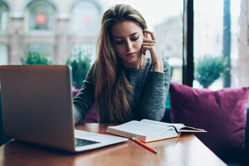 Trendy thoughtful hipster girl spending time for reading literature and studying on netbook at cafeteria, clever pensive female writer searching information for new article in laptop computer