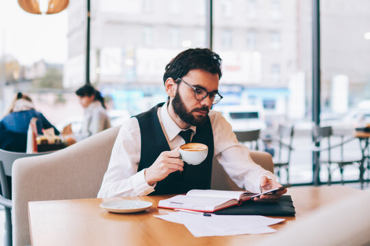 Caucasian hipster guy in trendy look reading sms message on modern smartphone from colleague while resting at cafeteria and enjoying cappuccino beverage, concept of technology and communication