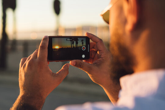 Close up back view of male holding mobile phone for shooting beautiful landscape of sundown during vacation, hipster guy using modern smartphone and application taking image of nature during trip