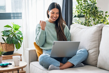 Pretty motivated woman celebrating something while working with laptop sitting on a couch at home