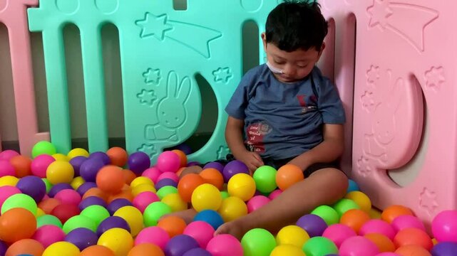 A child living with cerebral palsy is playing in a colorful ball pool in the occupational therapy room. A young child with a nasogastric tube sits among a colorful sea of plastic balls. 